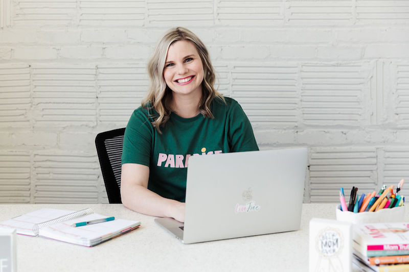 Blonde woman in a green shirt at a desk behind a laptop with a white wall.