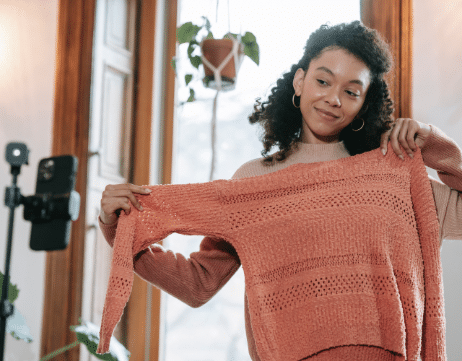 A woman with curly black hair and hoop earrings holds up a peach-colored sweater to herself